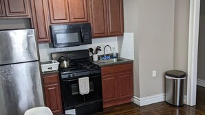 Cucina compatta con mobili in legno, frigorifero in acciaio inossidabile, piano cottura nero e forno a microonde. Nell'area soggiorno, si trova un tavolino con una sedia, un cestino e un divano.