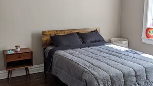 Una camera da letto con una testiera in legno, una coperta grigia, un comodino con una lampada e una finestra vista esterna.