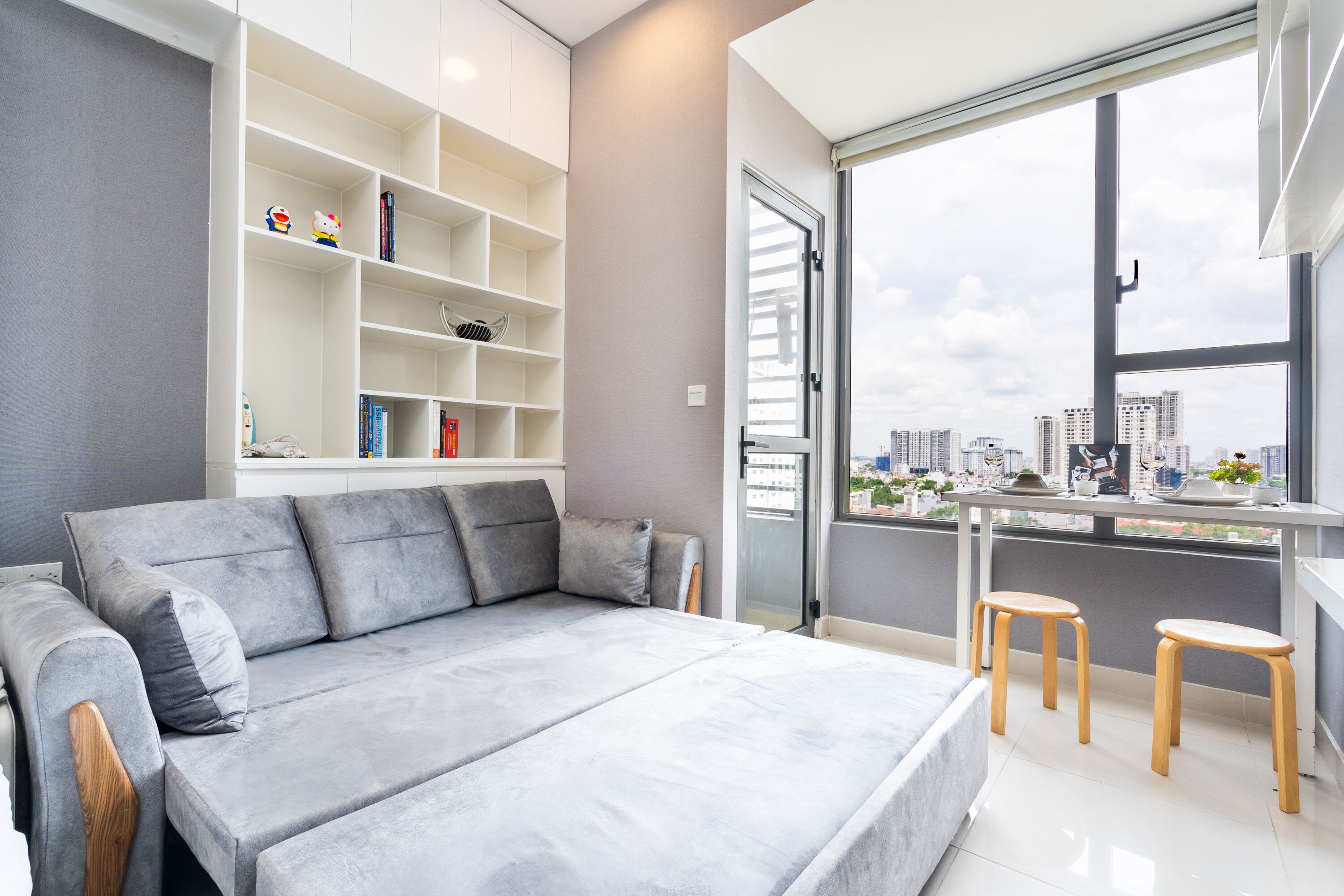 Ein modernes Schlafzimmer mit grauem Sofa, weißem Schreibtisch mit zwei Hocker und einem großen Fenster mit Blick auf die Stadt.