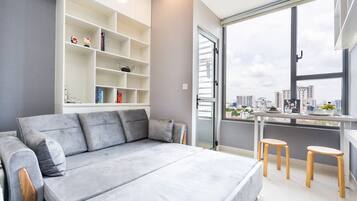 Ein modernes Schlafzimmer mit grauem Sofa, weißem Schreibtisch mit zwei Hocker und einem großen Fenster mit Blick auf die Stadt.