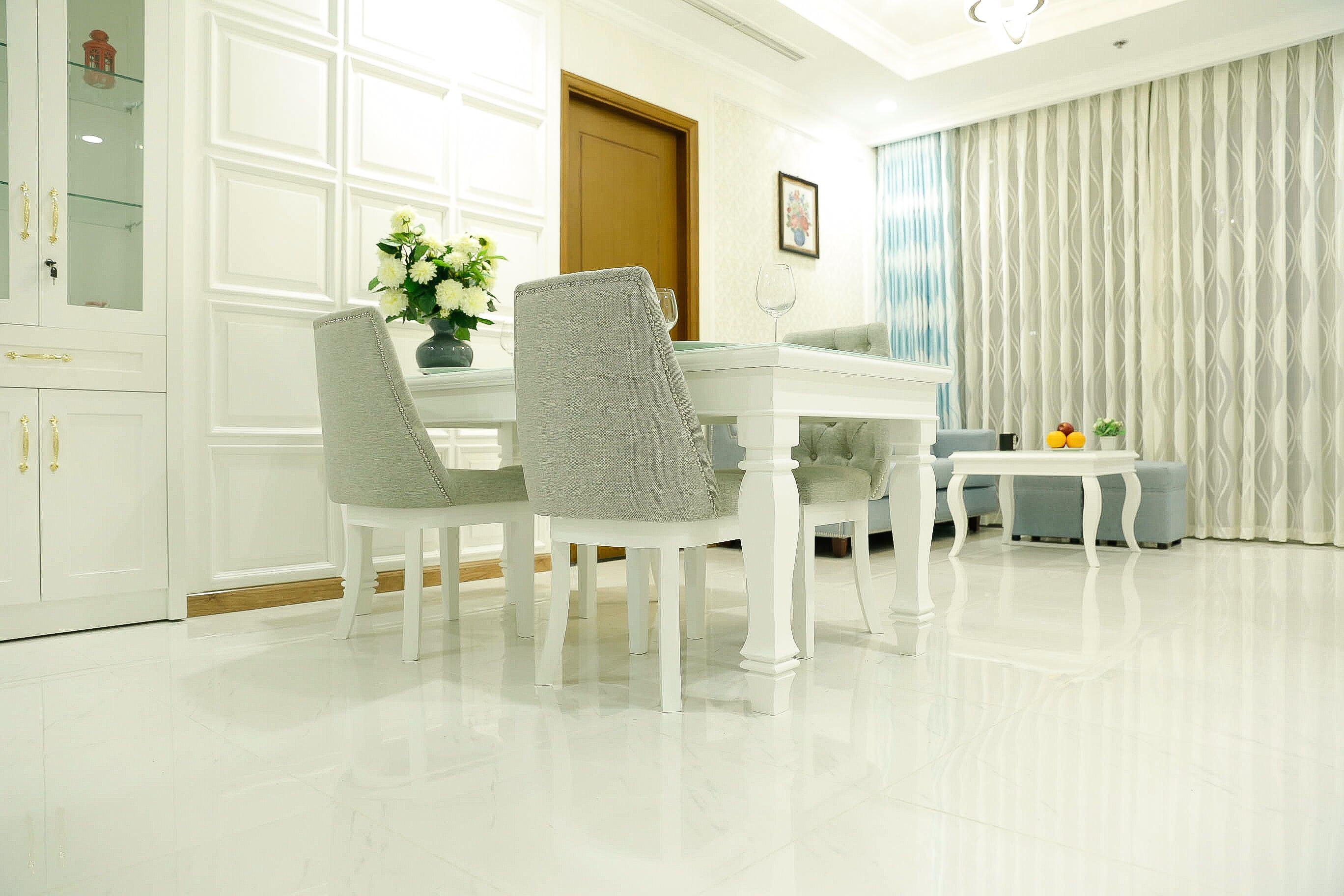 A dining area with a white table, two grey upholstered chairs, a vase with flowers, and a glass of wine on the table.