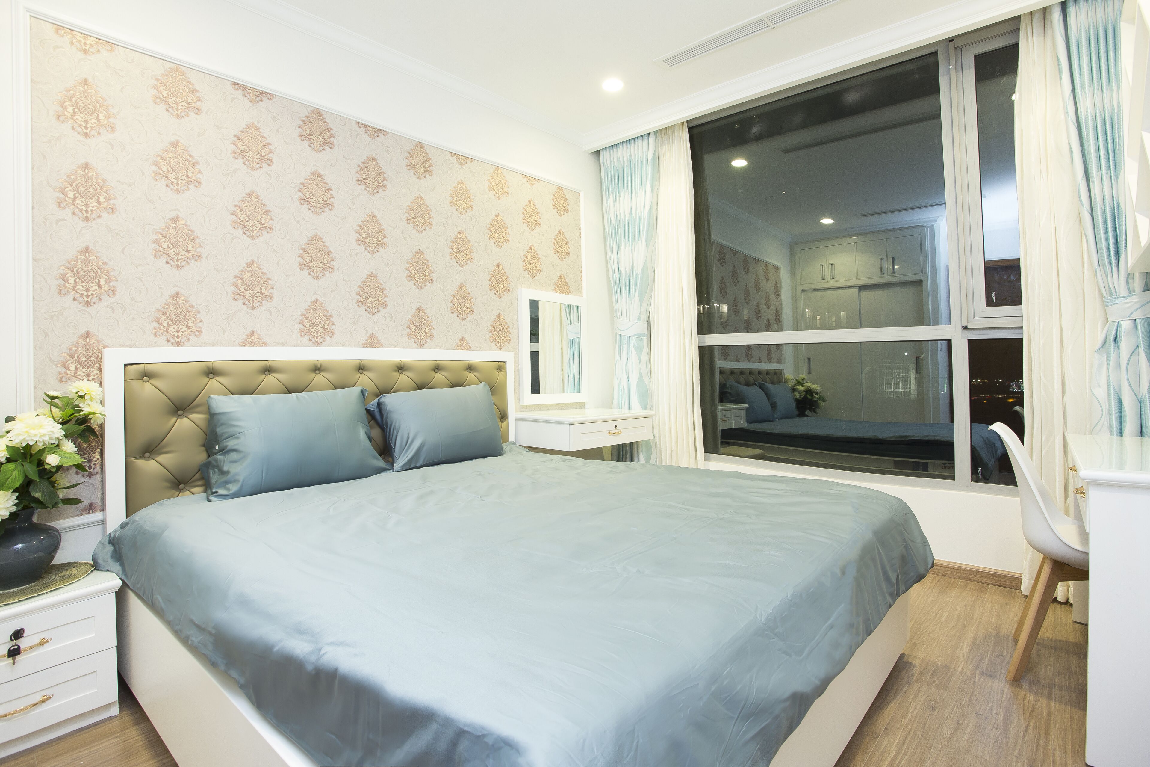 Un dormitorio con una cama grande, una mesita de noche con un florero y vistas a la ciudad desde una ventana.