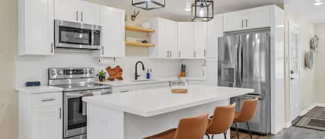 A modern kitchen with white cabinetry, stainless steel appliances, a central island with bar stools, and pendant lighting.
