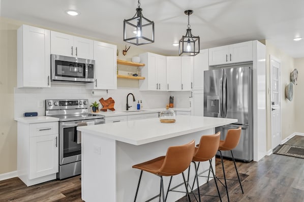 A modern kitchen with white cabinetry, stainless steel appliances, a central island with bar stools, and pendant lighting.