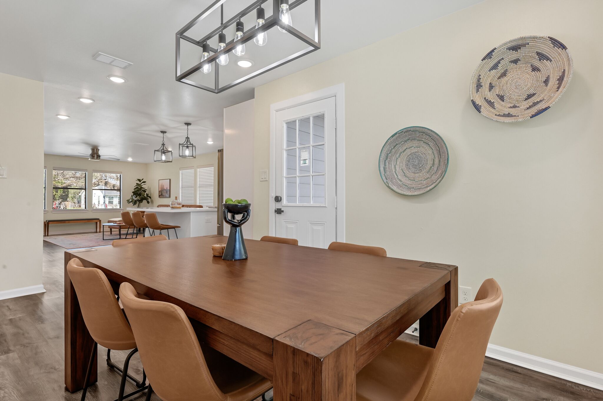A dining area with a large wooden table, six chairs, pendant lighting, and decorative plates on the wall.