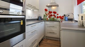 A modern kitchen with white cabinets, a stainless steel oven, a poppy flower decoration, and a window with a red blind. - Albies End (Battle)