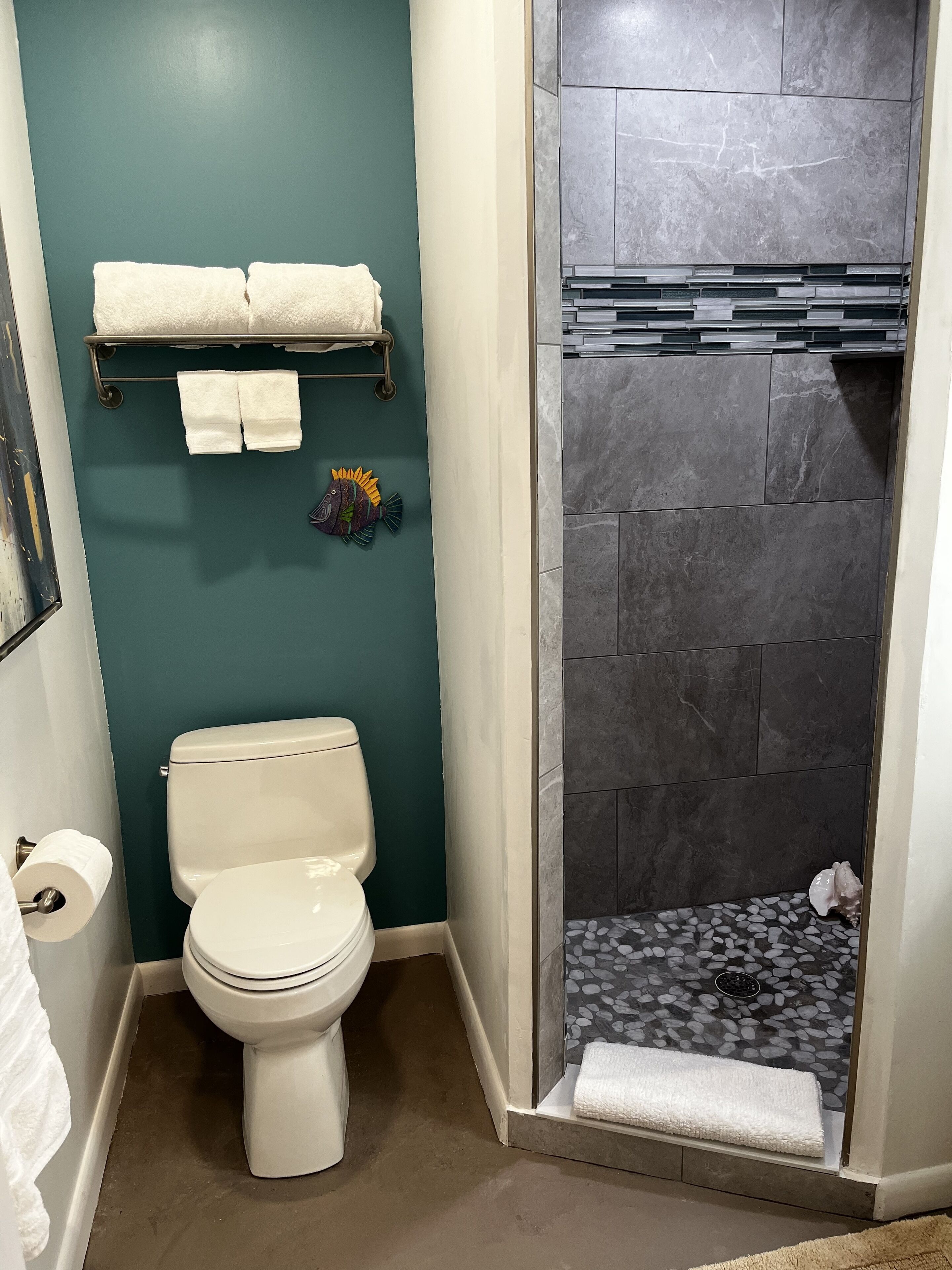 A bathroom with a white toilet, teal walls, a shower with stone tiles, and a towel rack with two towels.