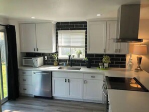 Uma cozinha moderna com armários brancos, painel de fundo preto, janela com persianas, micro-ondas, pia e fogão.