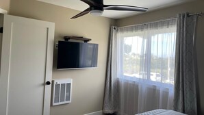 Camera da letto con ventilatore a soffitto, televisore a parete, finestra con tende leggere e termosifone.