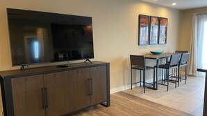 Un soggiorno moderno con una grande TV a schermo piatto su un mobile in legno, un'area pranzo con tavolo e sedie, e due opere d'arte incorniciate appese al muro.