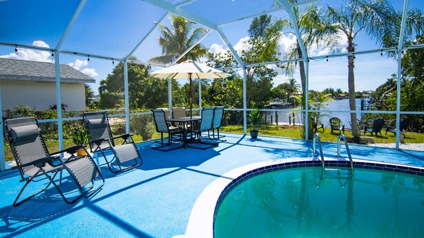 A covered outdoor swimming pool with lounge chairs and a table, surrounded by palm trees and a clear blue sky. - Ellie’s Landing 1 – Chill & Grill Dockside Oasis (Port Charlotte)