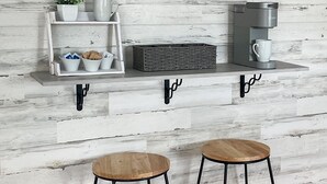 A kitchen with a rustic wooden wall, a coffee machine, two stools, and a shelf with various items including a plant and cups. - Ellie’s Landing 1 – Chill & Grill Dockside Oasis (Port Charlotte)