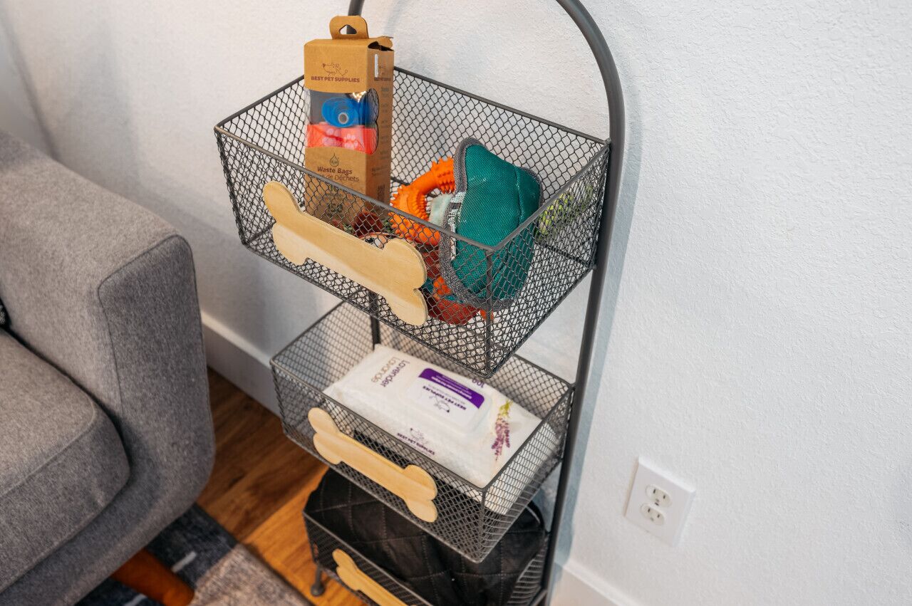 Een zwarte draadmand met een houten handvat, gevuld met een doos met de tekst 'BEST PET SUPPLIES', een groen kussen en een witte doos met een paars etiket.