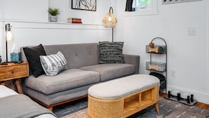 En moderne stue med en grå sofa, en træbænk, en gulvlampe og en vægmonteret hylde med bøger og en plante.