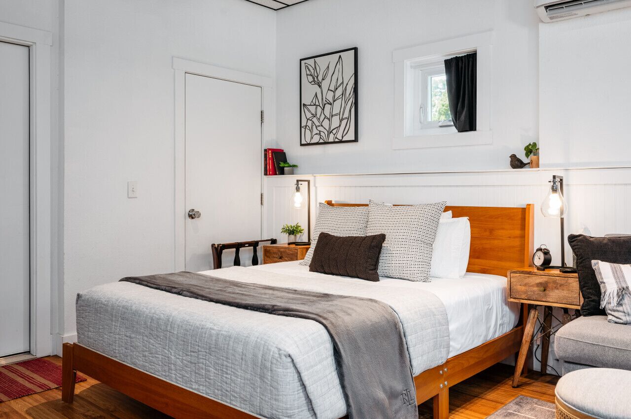 Un dormitorio ordenado con una cama de madera, una mesita de noche con lámpara y un ventanal con cortinas.