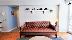 A brown leather sofa in a room with a wooden floor, a white wall, and a shelf with various items. - 2D Apartment Impeccable - Air Conditioned - Metro Los Héroes (Santiago)