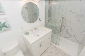 A bathroom with a white vanity, a round mirror, a glass shower enclosure, and a herringbone-patterned floor.