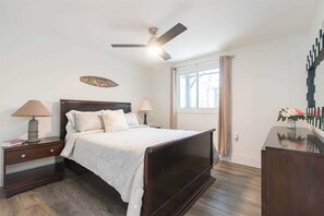A bedroom with a dark wooden bed, two bedside tables with lamps, a ceiling fan, a window with curtains, and a decorative wall piece.