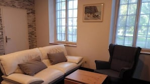 Una habitación con un sofá de cuero blanco, una mesita de madera, un sillón oscuro y un ventanal con un cuadro.