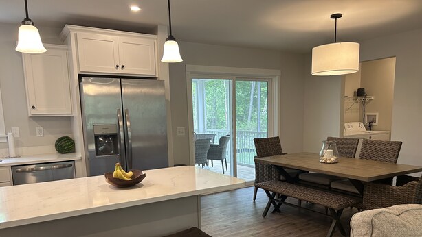 A modern kitchen with white cabinets, a stainless steel refrigerator, a wooden dining table with wicker chairs, and pendant lights.