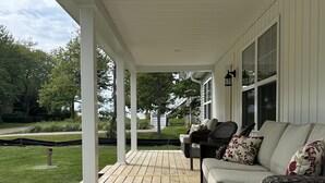A covered porch with a wooden deck, a wicker sofa with cushions, and a view of a green lawn and trees.
