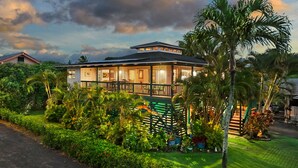 Une maison à deux étages dotée d’un balcon, entourée d’une végétation luxuriante et de palmiers.