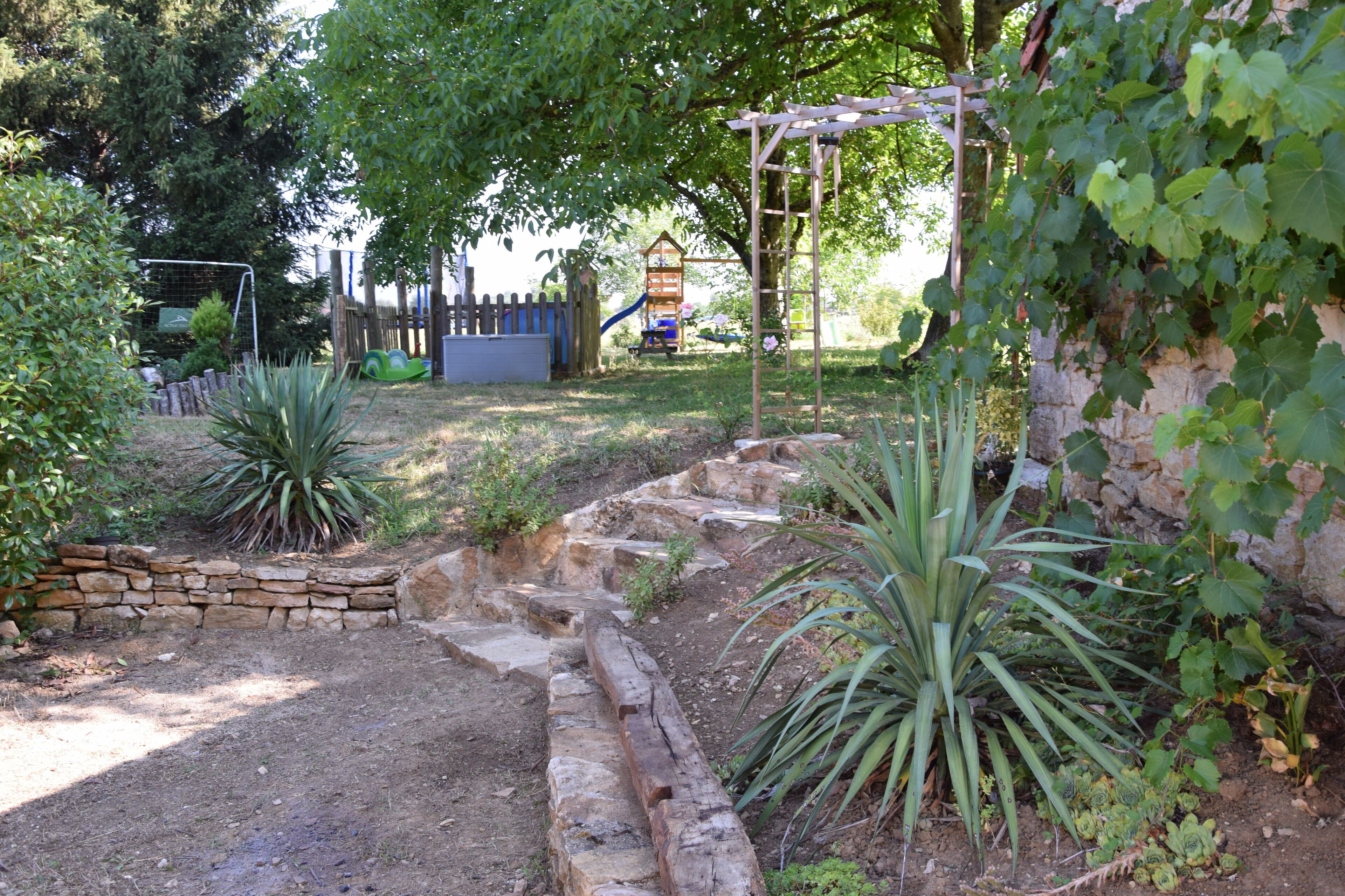 A garden with a stone wall, a wooden pergola, and a playground in the background.