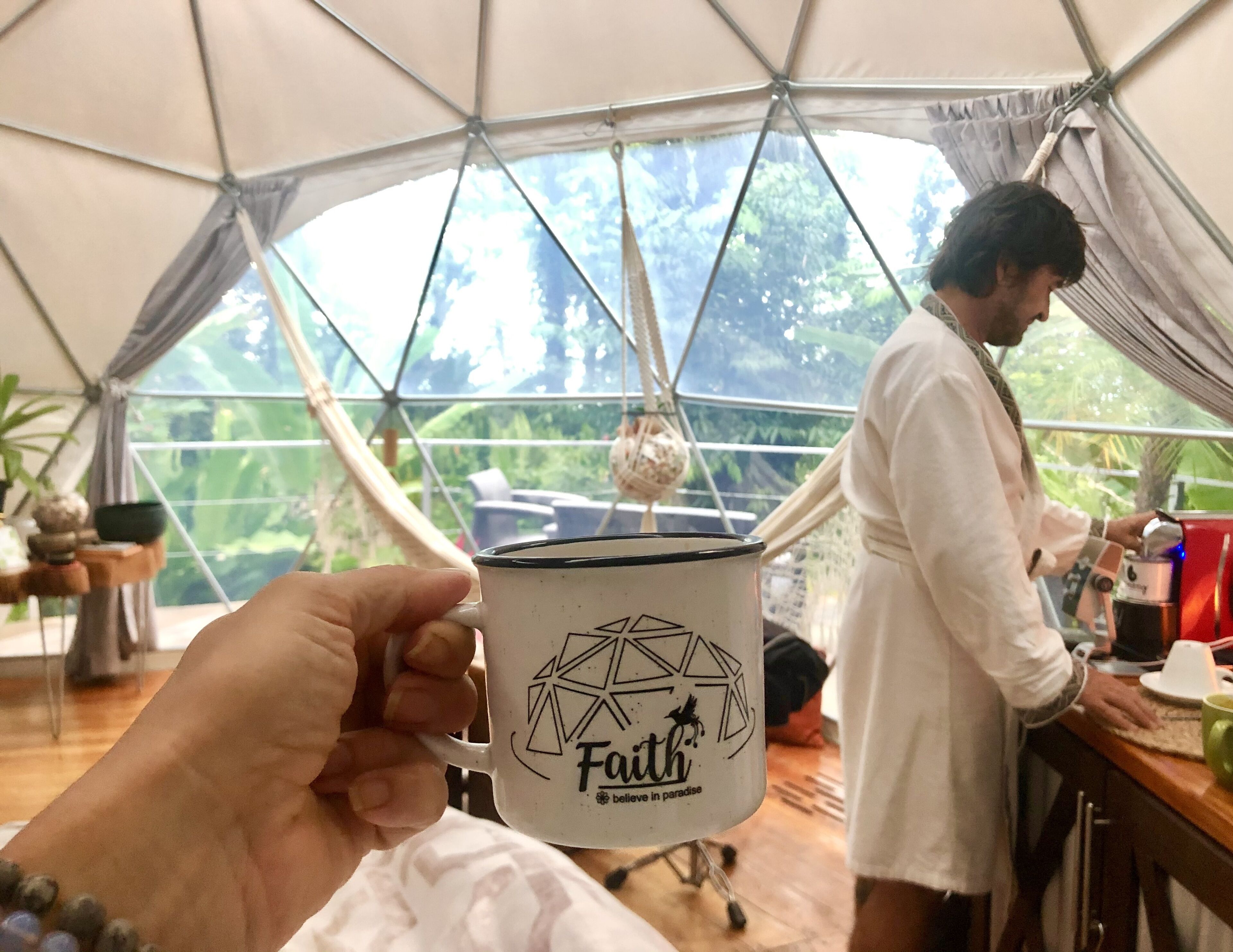 A person holding a mug with a geometric design and the word "Faith" on it. In the background, there is a tent with a view of greenery outside.