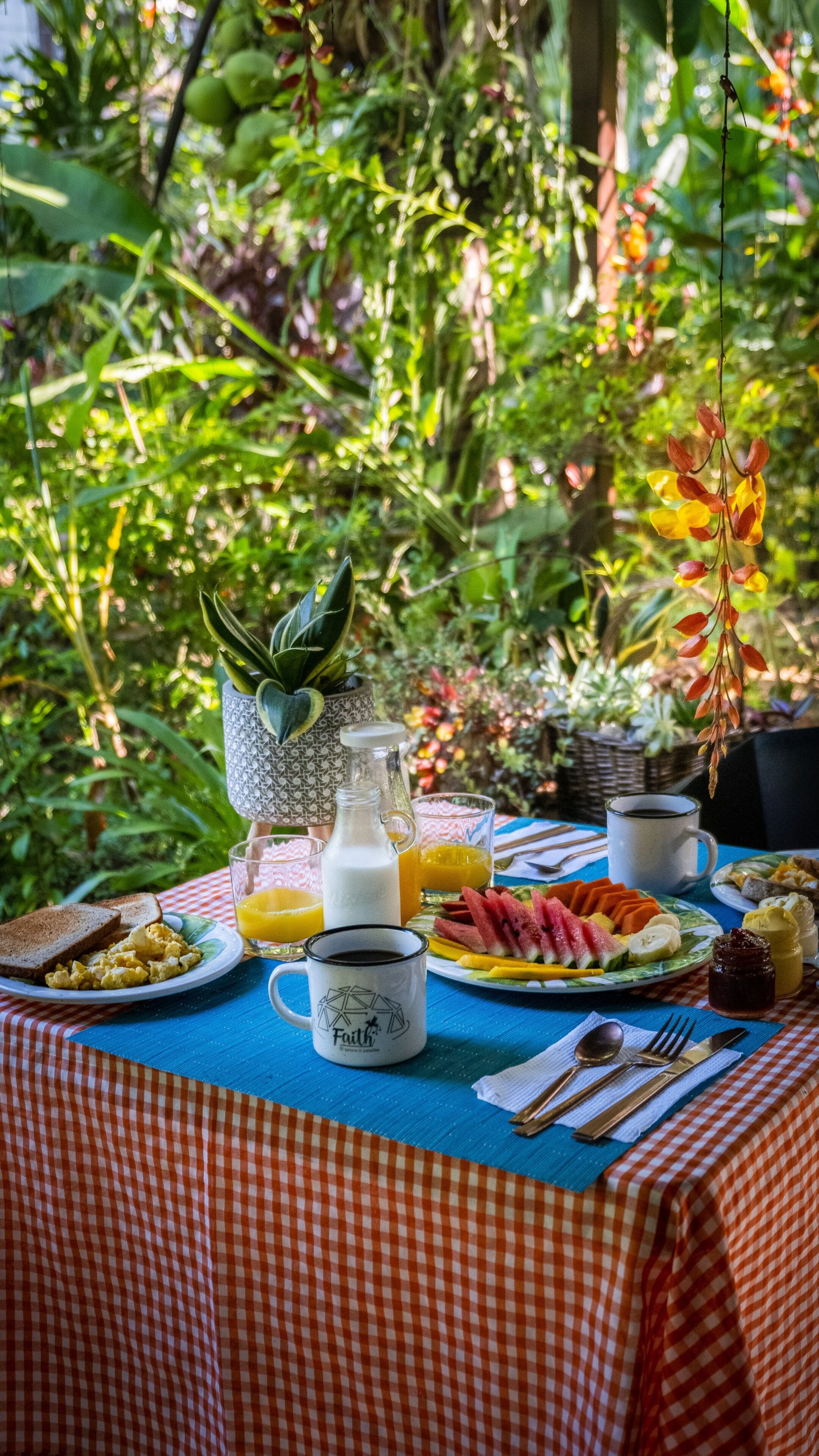 A breakfast table set outdoors with a checkered tablecloth, plates of food, a pitcher of milk, a glass of orange juice, and a mug.