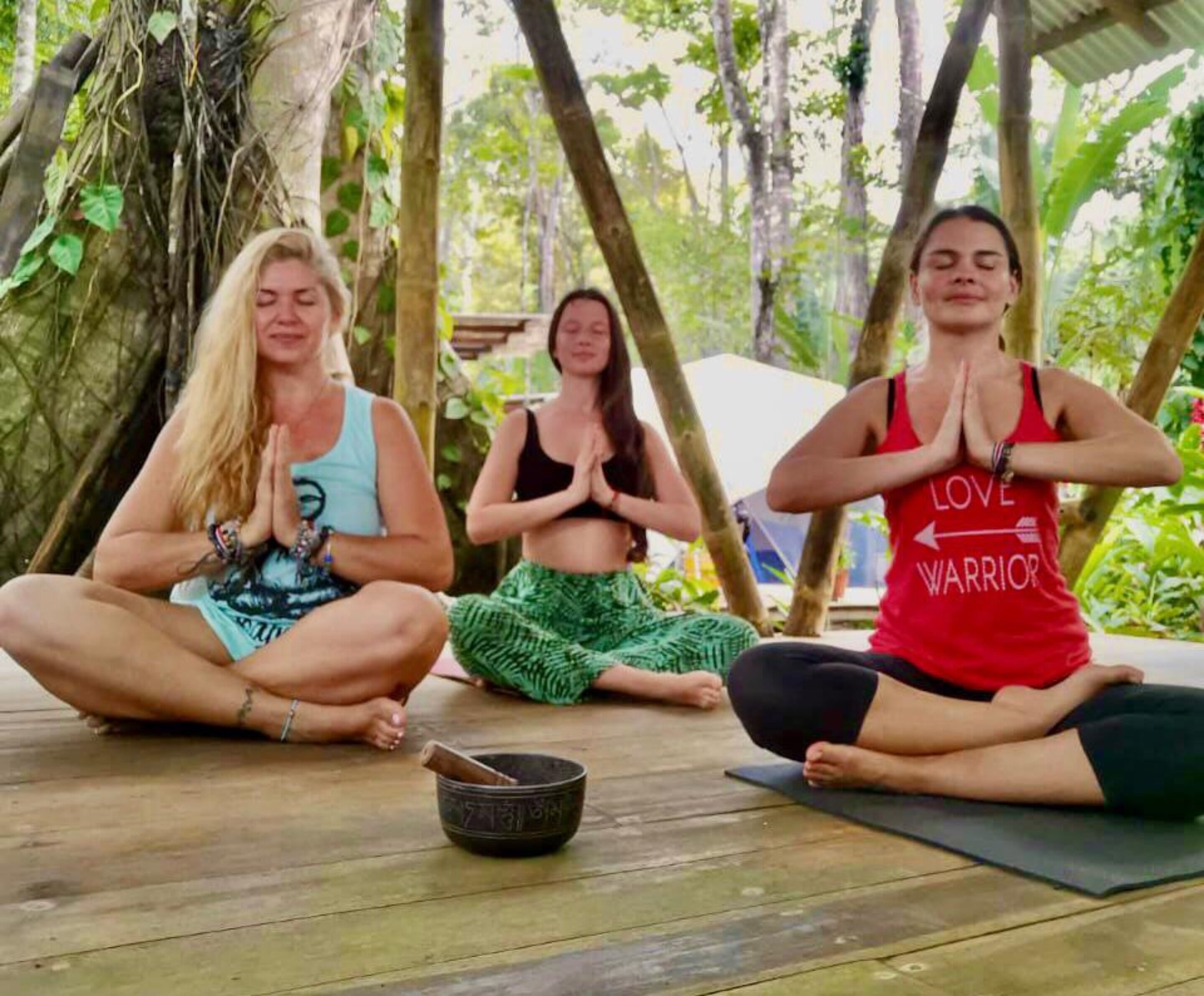 Three individuals meditating outdoors on a wooden deck with a gong nearby.