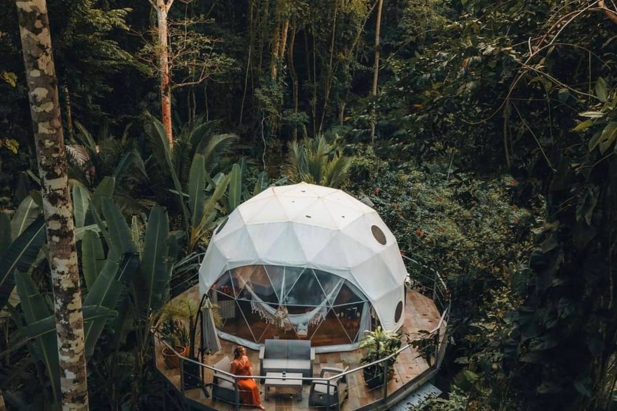 A geodesic dome structure surrounded by dense tropical foliage, featuring a wooden deck and outdoor seating area.