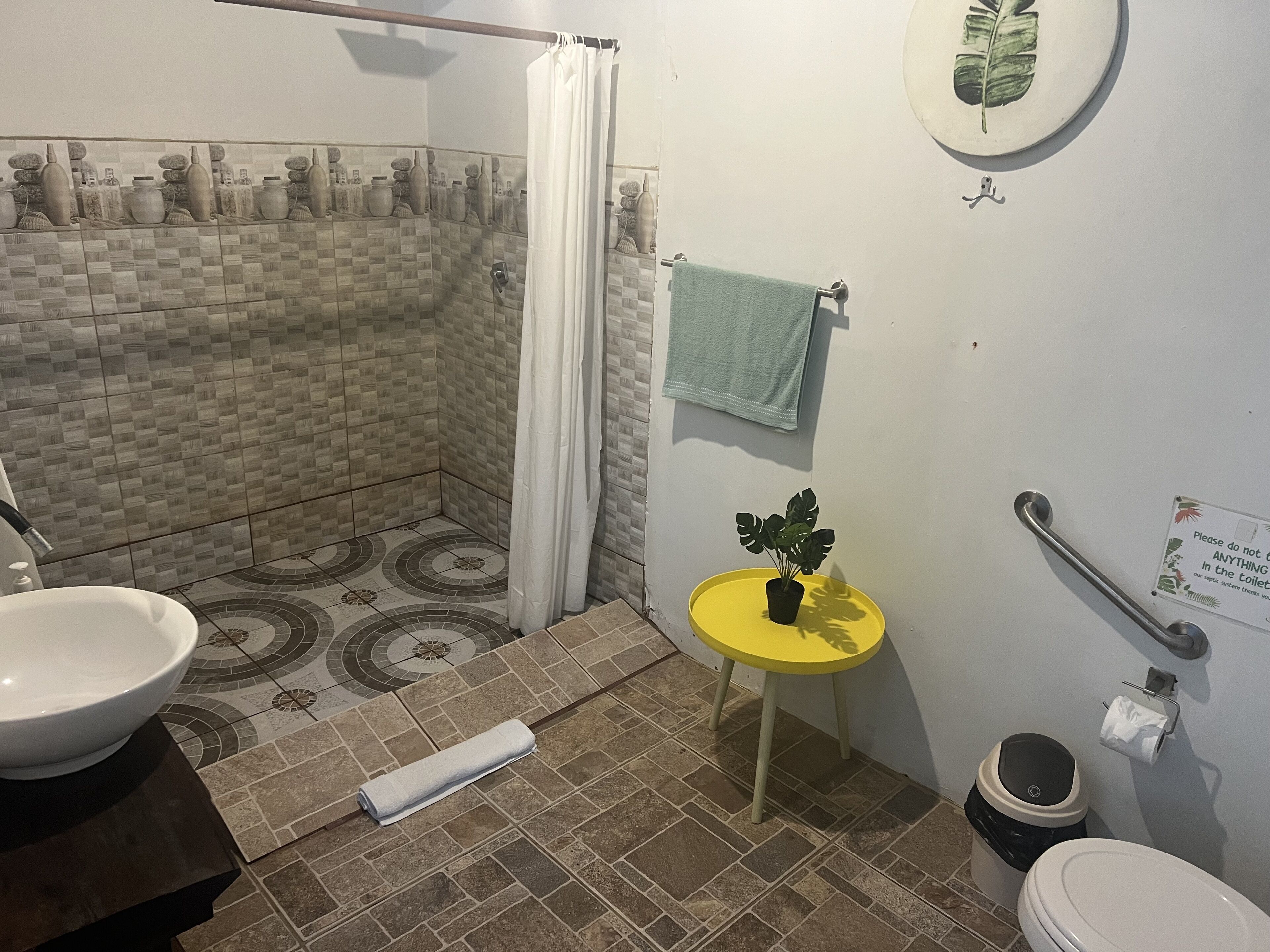 Une salle de bain équipée d’une douche, d’un lavabo, de toilettes et d’une petite table sur laquelle est posée une plante.