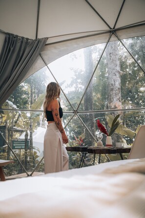 A person standing in a room with a large glass window, looking out at a tropical garden. - Faith Glamping Dome Costa Rica (Cahuita)