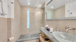 Un bagno moderno con piastrelle color crema, cabina doccia in vetro, lavabo con specchio e un piccolo gradino che conduce alla doccia.