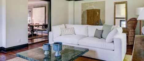 A living room with a white sofa, a glass coffee table with two blue vases, a dining area with a wooden table and chairs, and a large mirror on the wall.