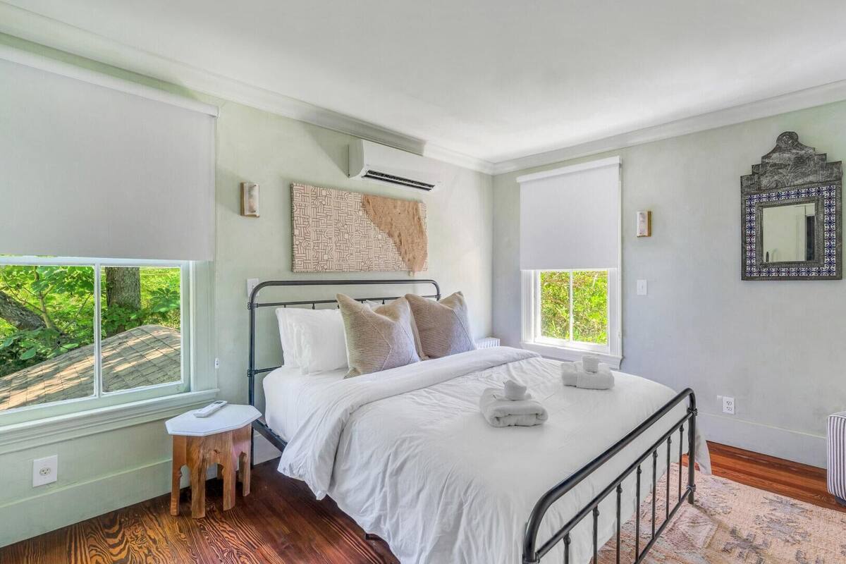 Quarto com cama, duas janelas, radiador, um banquinho pequeno e um espelho na parede.