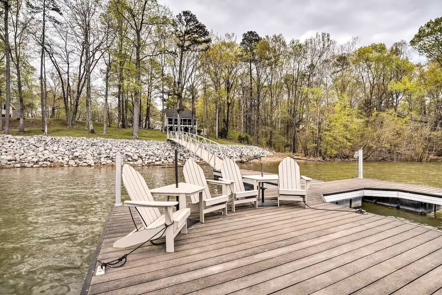 A wooden deck with Adirondack chairs and a table, overlooking a river with a rocky shoreline and trees in the background.