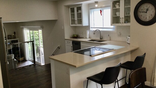 A modern kitchen with an island, bar stools, a window with a red hanging ornament, and a clock on the wall.