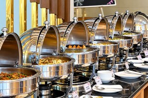 Tarjolla on runsas buffet, jossa on useita ruostumattomasta teräksestä valmistettuja lämpöastioita, joissa on erilaisia ruokia. Astiat on aseteltu tiskille, ja niiden yhteydessä on tarjoiluastioita ja ruokalappuja.