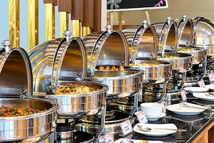 A buffet with multiple stainless steel chafing dishes containing various foods, arranged on a counter with serving utensils and labels.