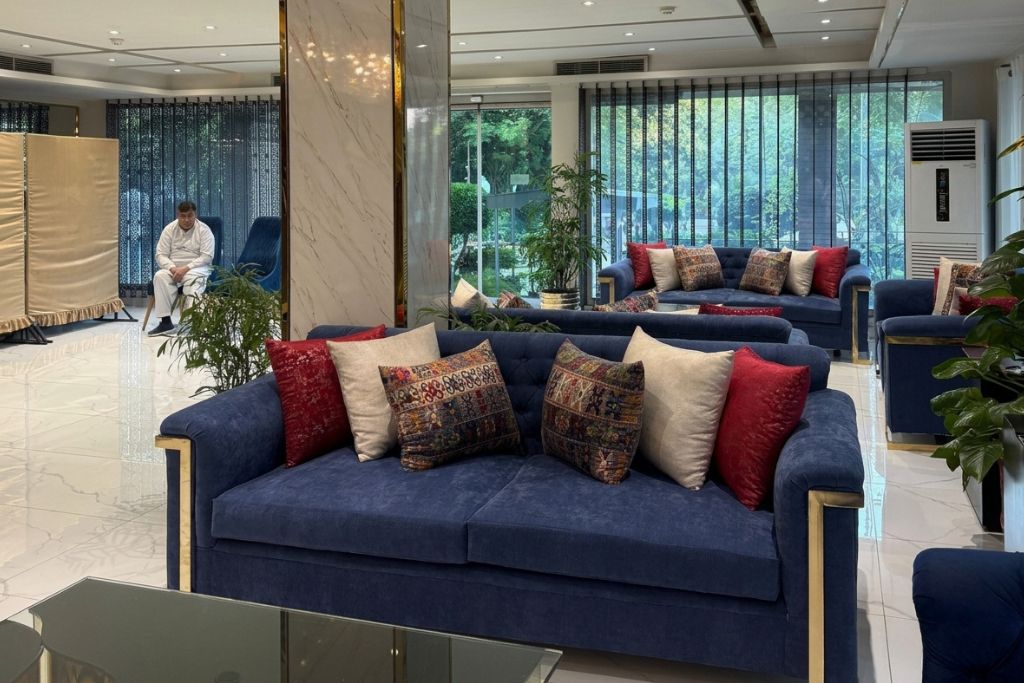 A modern living room with a blue sofa set, a glass coffee table, and a large window with a view of greenery.