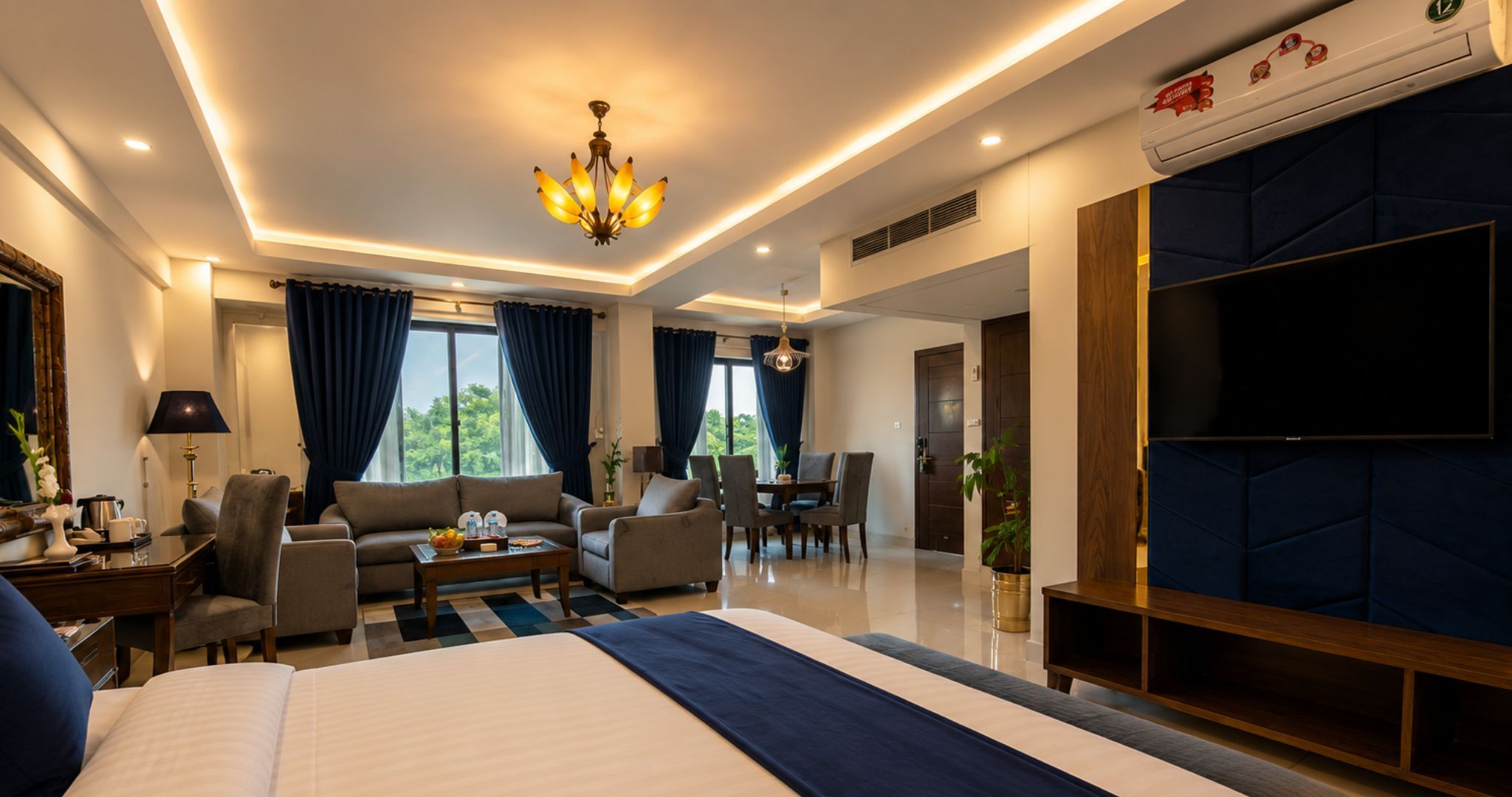 Ein modernes Hotelzimmer mit einem großen Bett, einer Sitzecke mit Sofas und einem Esstisch, einem Fernseher und Blick ins Freie.