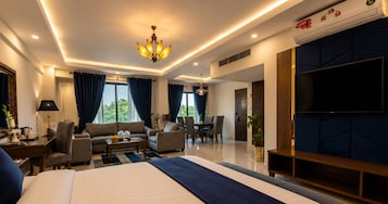Una habitación de hotel moderna con una cama grande, una zona de estar con sofás y una mesa de comedor, un televisor y vistas al exterior.
