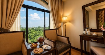 Habitación de hotel con un ventanal grande, una mesa de comedor con tarta y una cesta de fruta, una lámpara y un espejo.