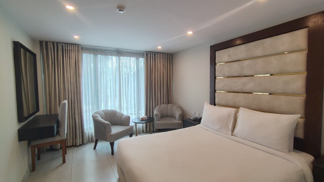 Habitació d'hotel amb un llit gran, dos seients, una taula petita, televisió i una finestra ampla amb cortines.
