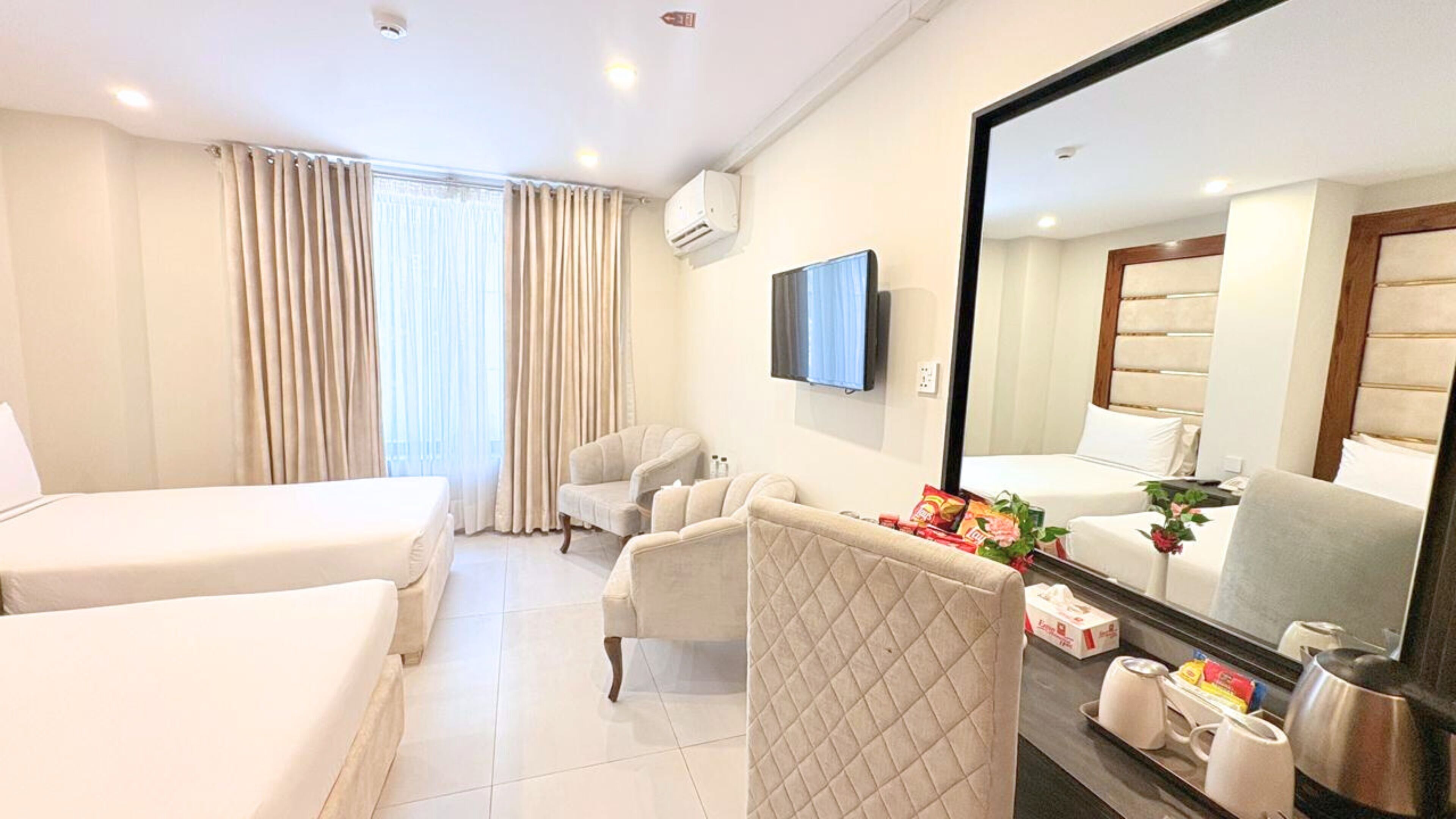 Habitación de hotel con dos camas, una zona de estar con una silla, un televisor de pantalla plana, un espejo grande y una mesita de noche con artículos de aseo.