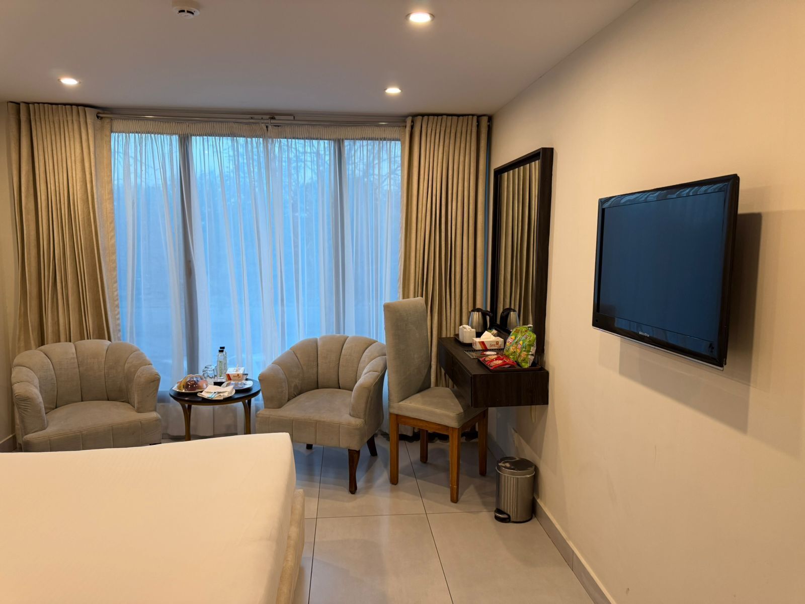 Habitación de hotel con cama, dos sillones, una mesita con una botella y vasos, un escritorio con una tetera y una bolsa, un televisor en la pared y una ventana con cortinas.