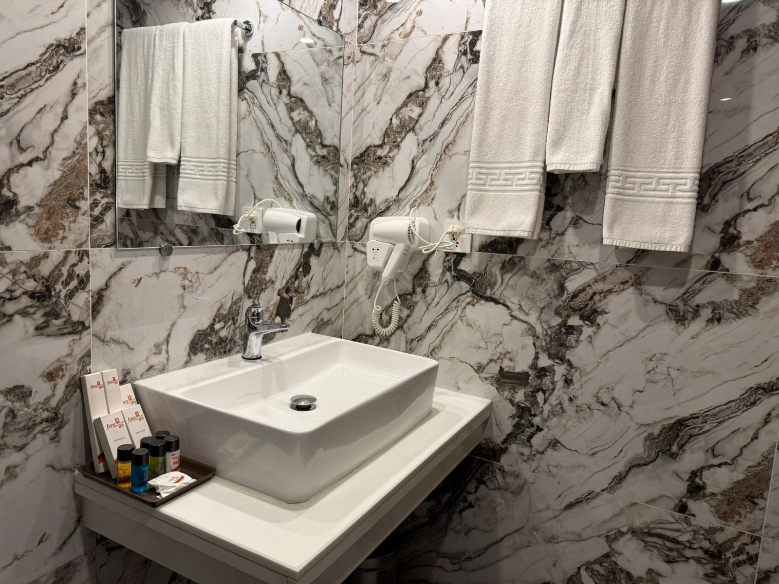 Un baño con lavamanos blanco, paredes de mármol y un espejo. Hay toallas colgadas en la pared y un secador de pelo sobre el mostrador.