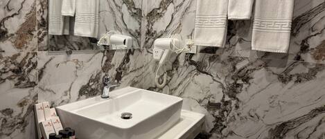 Un baño con lavamanos blanco, paredes de mármol y un espejo. Hay toallas colgadas en la pared y un secador de pelo sobre el mostrador.