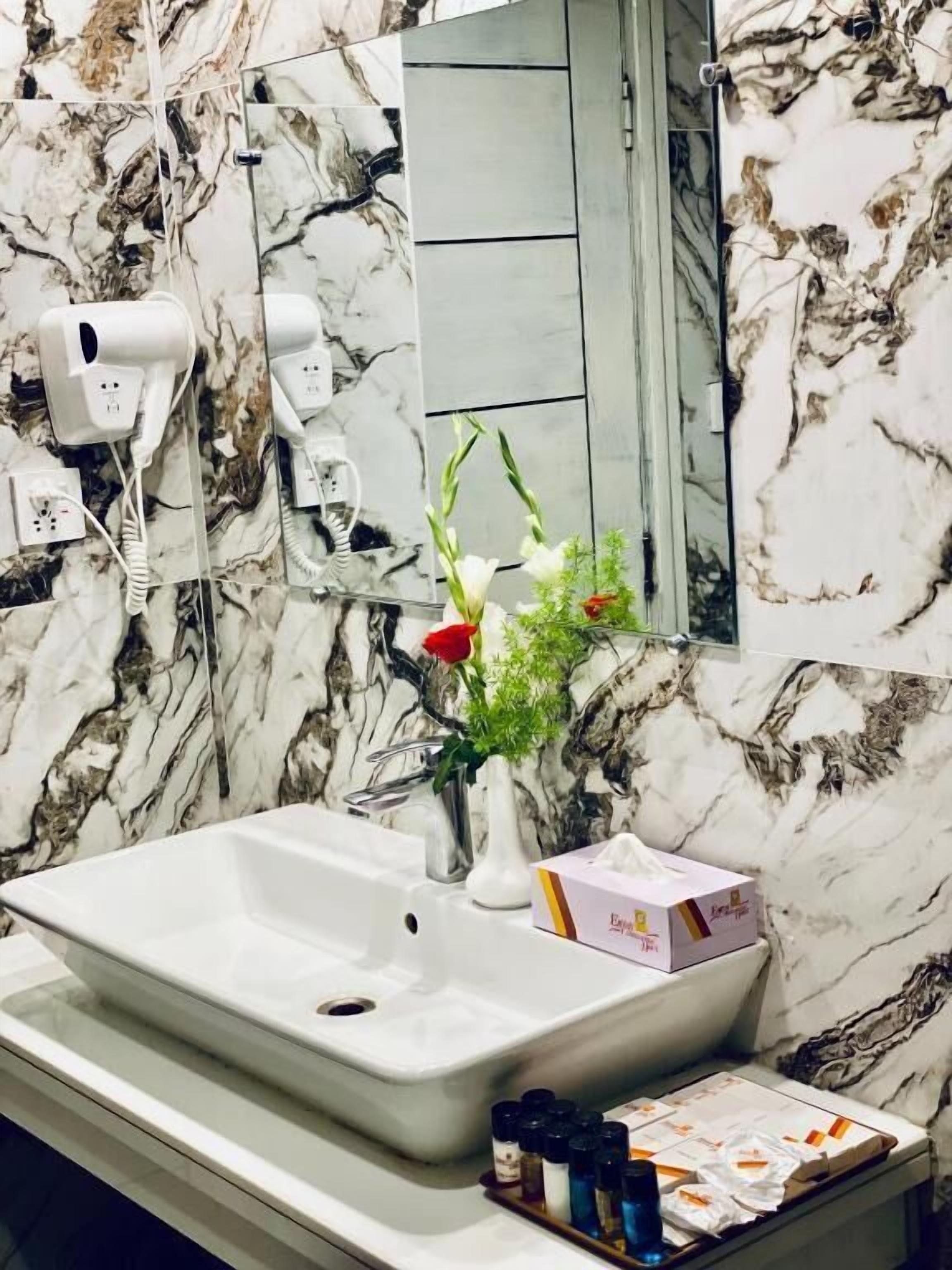 A bathroom with a marble backsplash, a white sink, a mirror, a vase with flowers, and a box of toiletries.